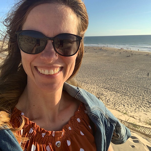 Frau mit schwarzer Sonnenbrielle und langen Haaren steht am Strand und lacht in die Kamera